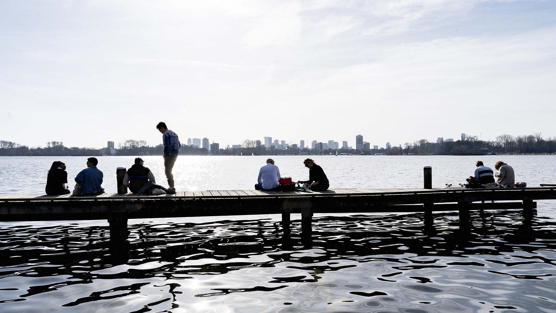 Mensen die op een steiger bij water zitten met skyline van Rotterdam op de achtergrond