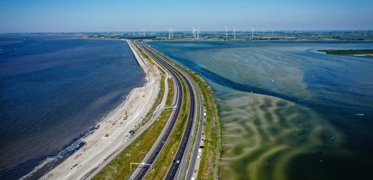 Luchtfoto van een weg op Afsluitdijk tussen Waddenzee en groen land met windmolens in de verte.