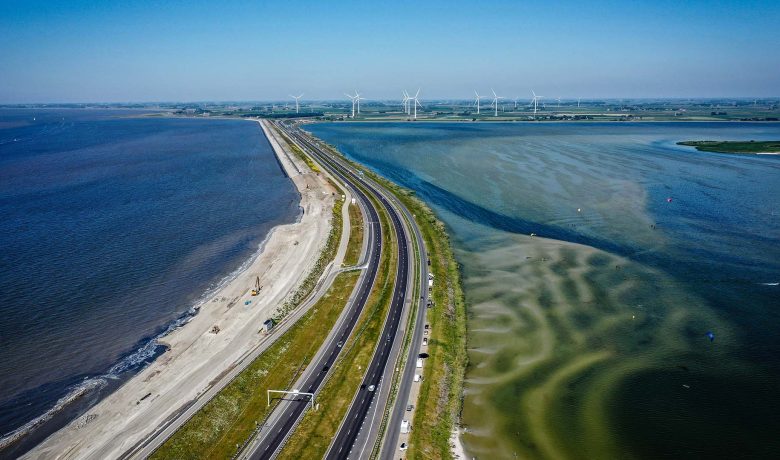 Luchtfoto van een weg op Afsluitdijk tussen Waddenzee en groen land met windmolens in de verte.