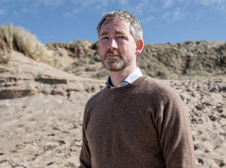 Alex Hekman, man met bruine trui staat op een zandduin onder blauwe lucht.