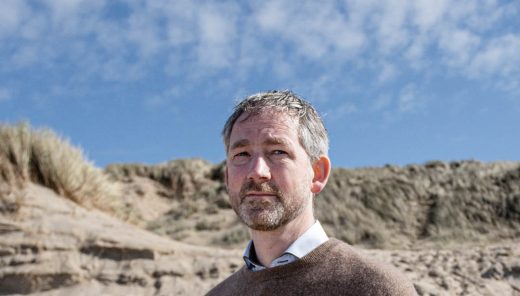 Alex Hekman, man met bruine trui staat op een zandduin onder blauwe lucht.