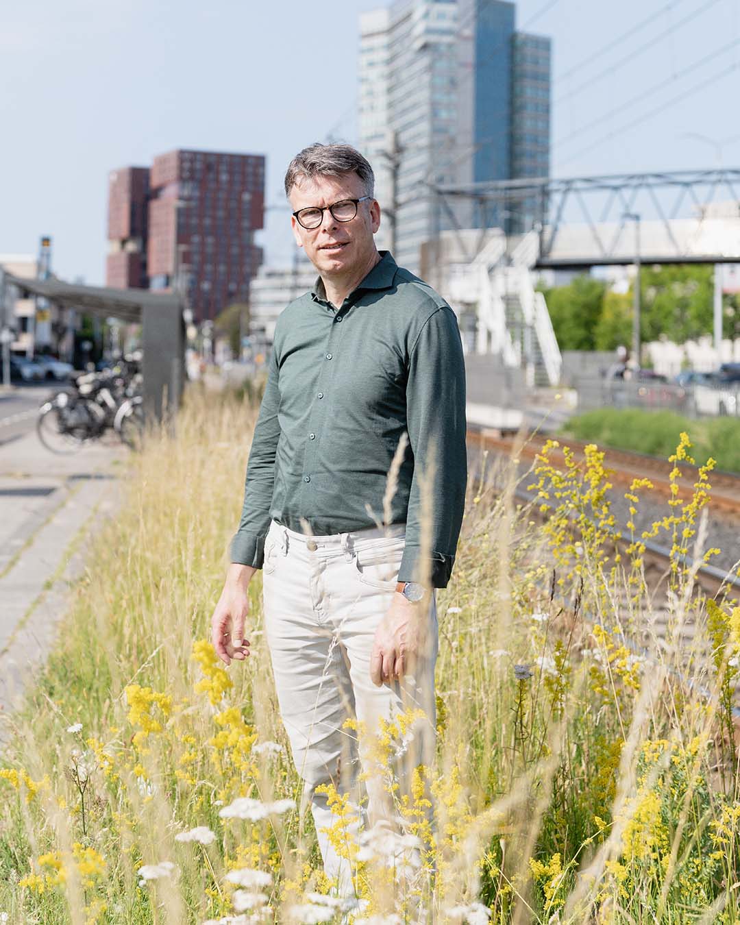Man in groen overhemd en witte broek, Henri Palm strategisch consultant verkeersmodellen bij Rijkswaterstaat, staat tussen wilde bloemen met gebouwen en spoorlijn op de achtergrond.