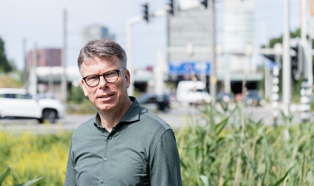 Man in groen overhemd, Henri Palm van Rijkswaterstaat, buiten bij een kruispunt met auto's en gebouwen op de achtergrond.