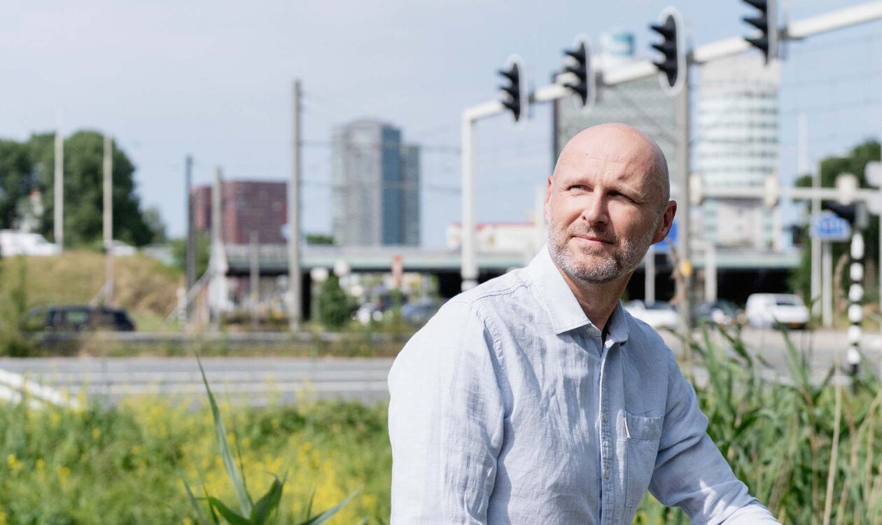 Man in wit overhemd, Ronald Plasmeijer expert verkeersmodellen bij Sweco, bij een verkeerslicht met moderne gebouwen op achtergrond.