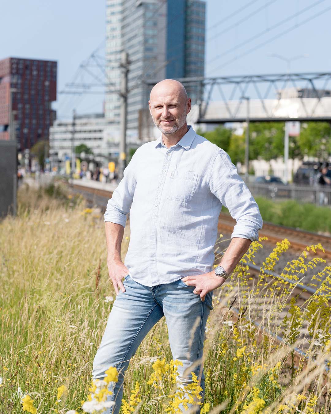 Man in witte blouse en jeans, Ronald Plasmeijer expert verkeersmodellen bij Sweco, poseert in een veld met hoge gebouwen en spoorweg op de achtergrond.