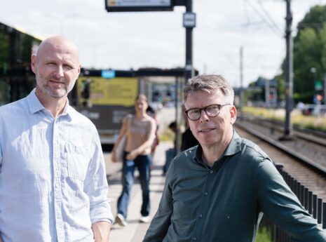 Twee mannen, Ronald Plasmeijer expert verkeersmodellen bij Sweco en Henri Palm strategisch consultant verkeersmodellen van Rijkswaterstaat, wachten bij tramhalte met tram op de achtergrond.