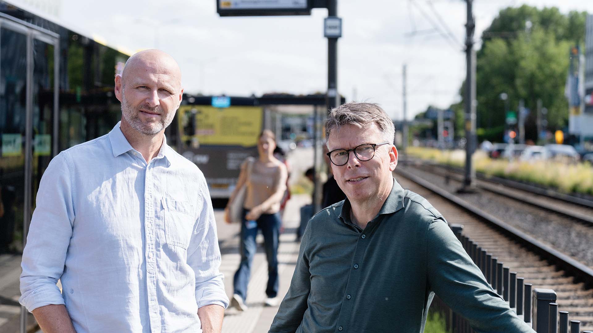 Twee mannen, Ronald Plasmeijer expert verkeersmodellen bij Sweco en Henri Palm strategisch consultant verkeersmodellen van Rijkswaterstaat, wachten bij tramhalte met tram op de achtergrond.