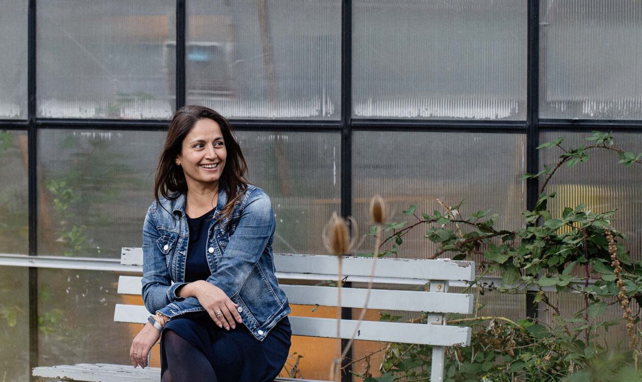 Sara Velhuizen, business unit manager Natuur en Klimaat bij Sweco, in een spijkerjack zit op een witte bank buiten bij een kas met planten op de achtergrond.