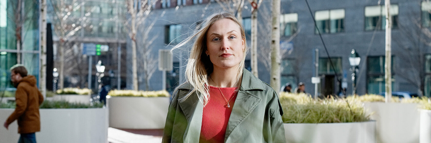 Sacha in groene jas en rode trui loopt buiten in Den Haag, gebouwen en planten op achtergrond.