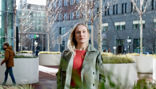 Sacha in groene jas en rode trui loopt buiten in Den Haag, gebouwen en planten op achtergrond.