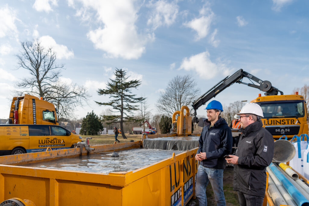 Twee mannen van VHGM met helmen bij bouwplaats met gele waterbeheer voertuigen en apparatuur.