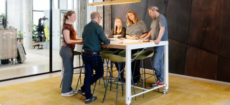 Samenwerking van meerdere collega's rondom een staande tafel. Ze kijken naar een technische bouwtekening en overleggen met elkaar.