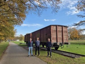 Drie mannen in groen landschap bij Westerbork voor treinspoor met goederenwagons