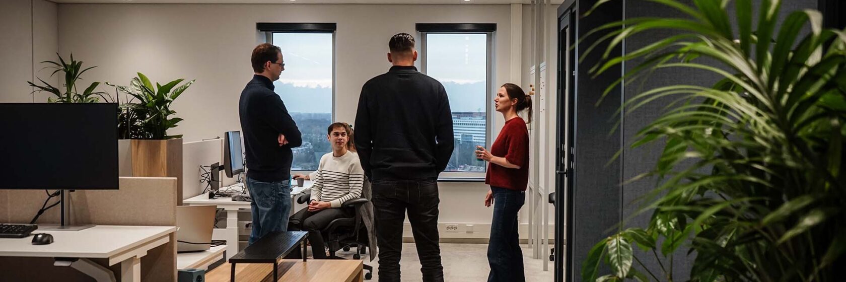 Vier mensen in gesprek binnen in lichte kantoorruimte met veel groene planten