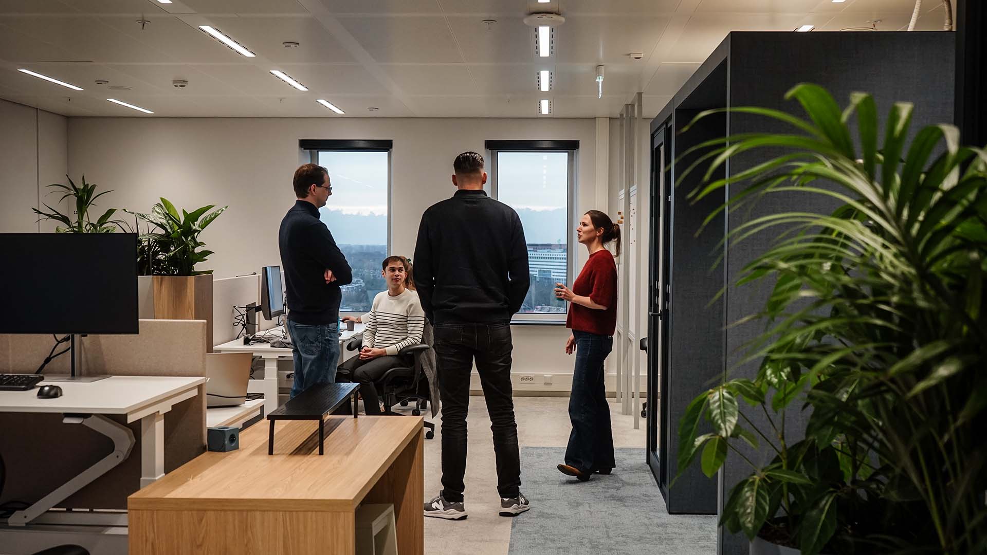 Vier mensen in gesprek binnen in lichte kantoorruimte met veel groene planten
