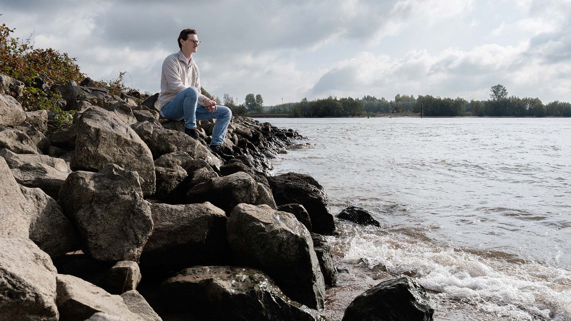 Man met bril, Maurits, op stenen oever bij Tiel-Waardenburg uitkijkend over rivier.