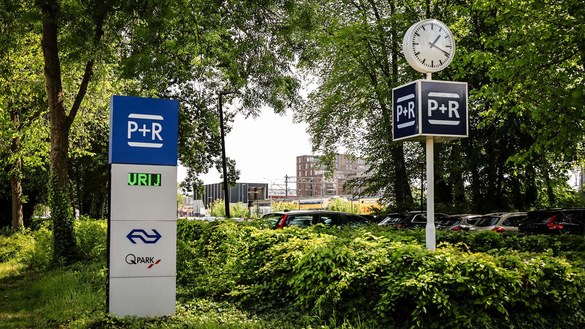 P+R bord bij station in groene omgeving met bomen en struiken met op achtergrond een hoog gebouw