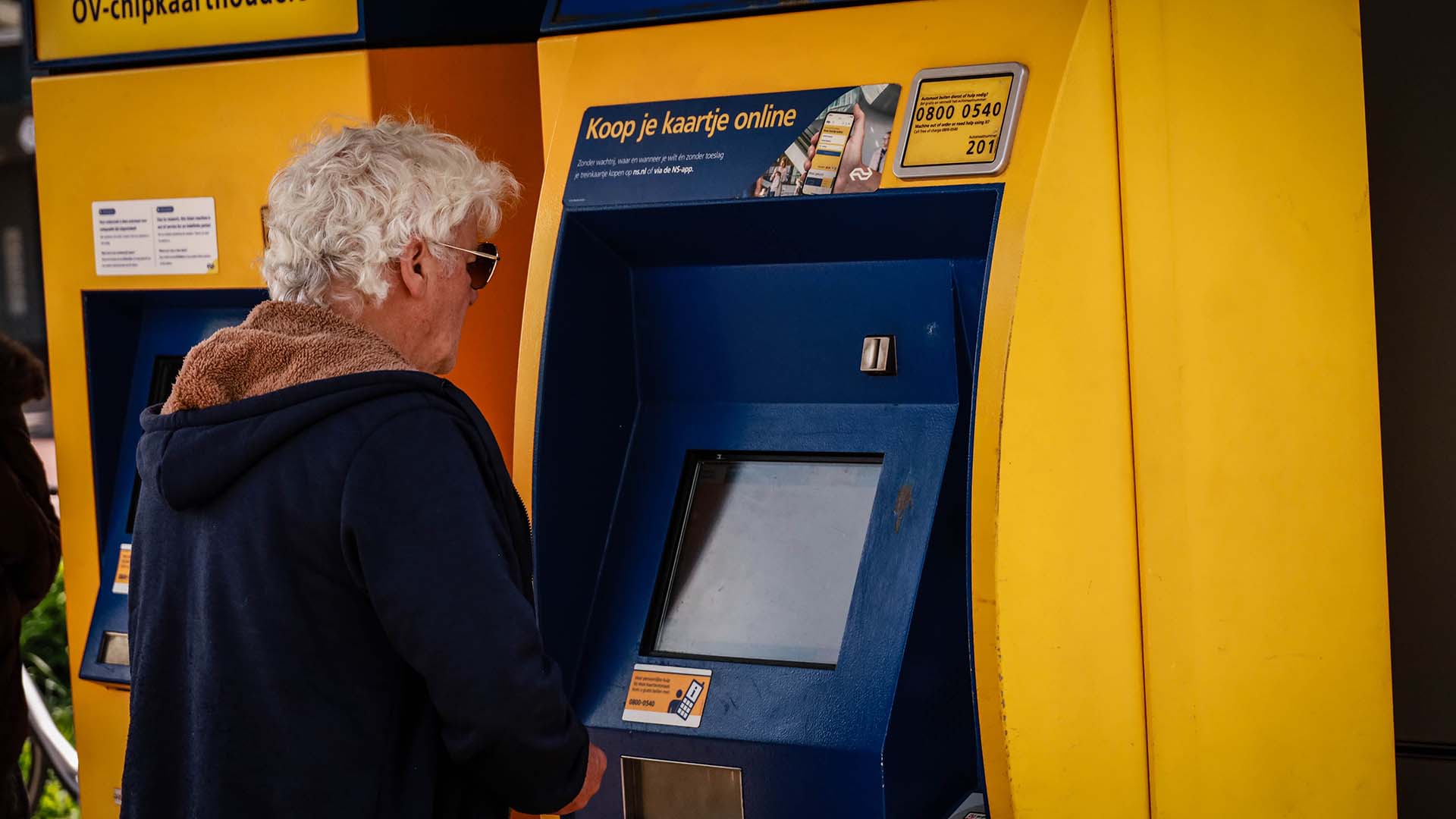 Oudere persoon bij geel blauwe kaartkoopautomaat op station