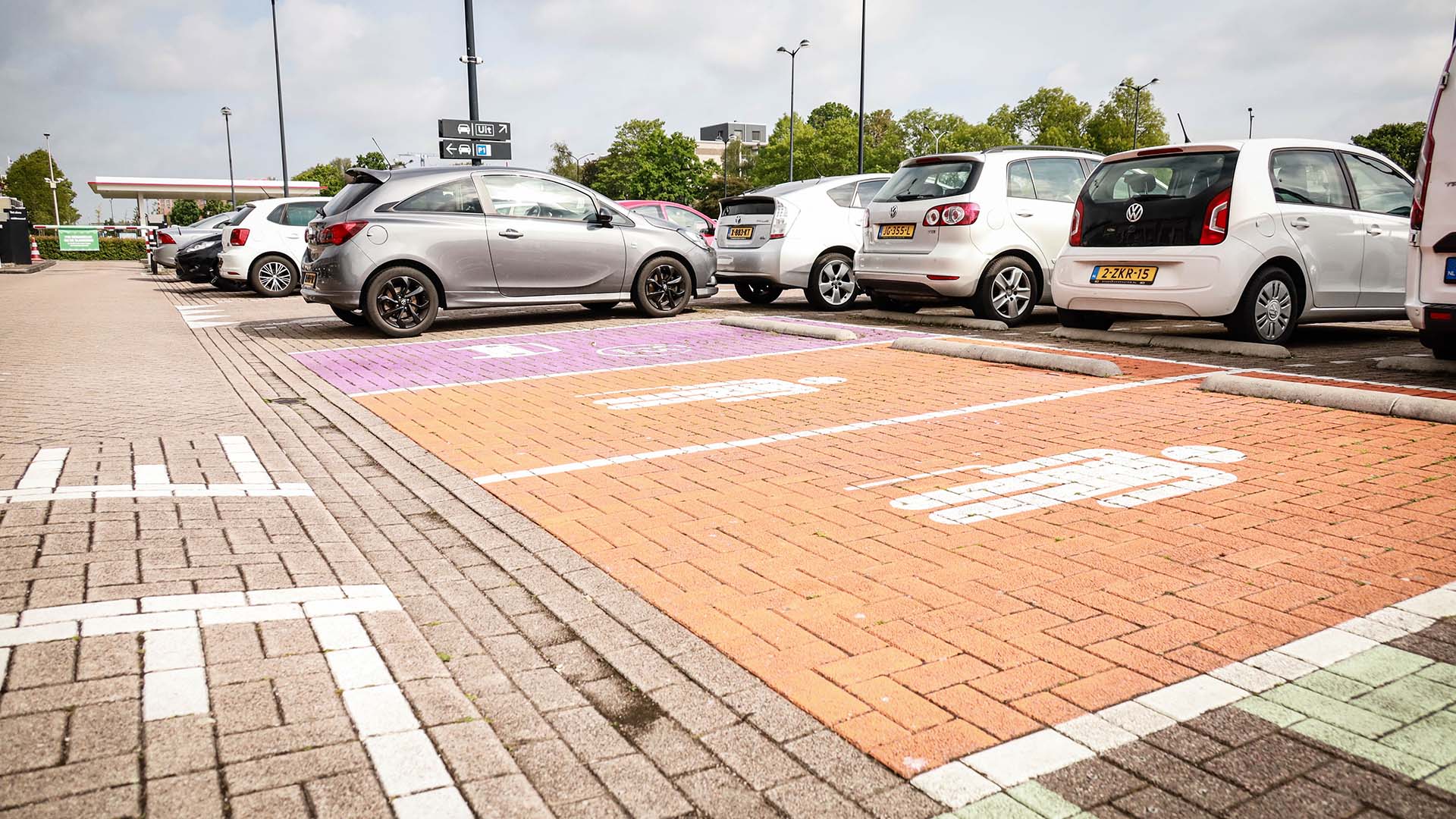 Oranje parkeervakken met pictogram voor minder validen en roze parkeervak voor ouders met kind op een parkeerterrein vol auto's.