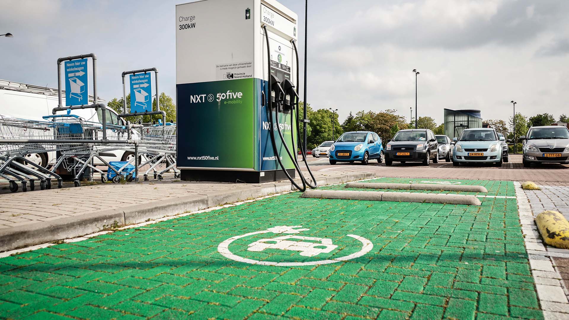 Groen parkeervak met oplaad icoon en oplaadstation ernaast. Geparkeerde auto's en winkelwagens op de achtergrond.