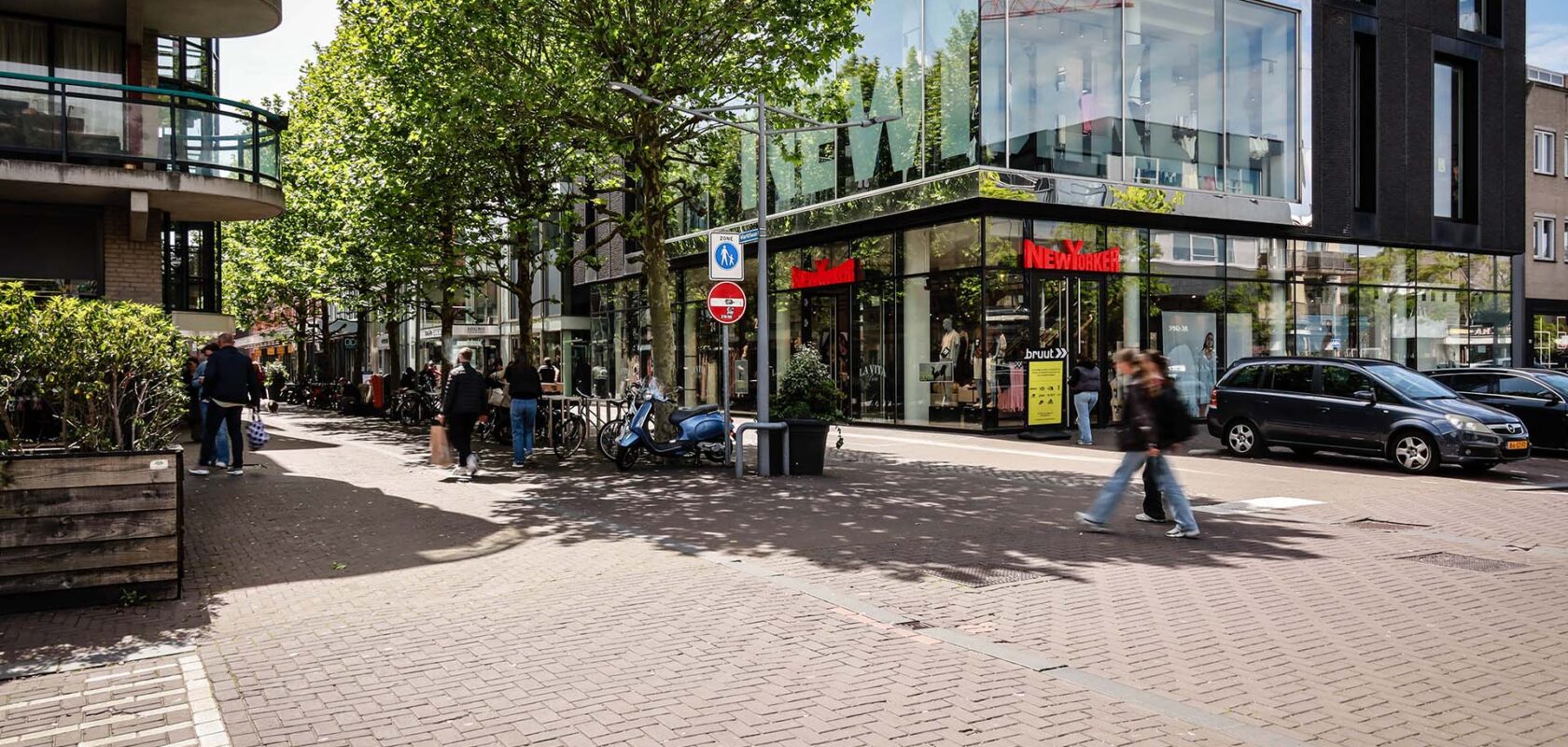 Straatbeeld winkelstraat met groene bomen, voetgangers en geparkeerde auto's.