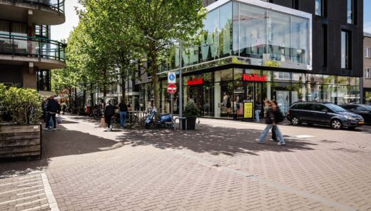Straatbeeld winkelstraat met groene bomen, voetgangers en geparkeerde auto's.