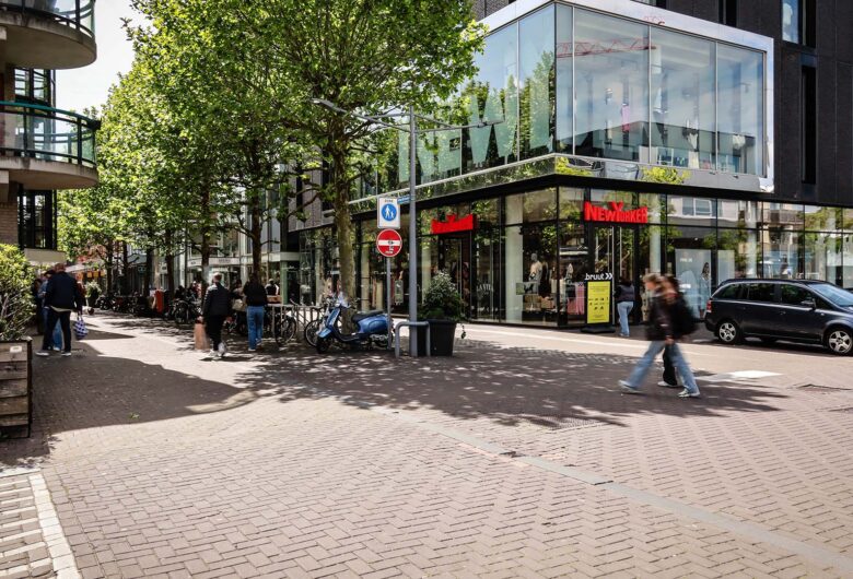 Straatbeeld winkelstraat met groene bomen, voetgangers en geparkeerde auto's.