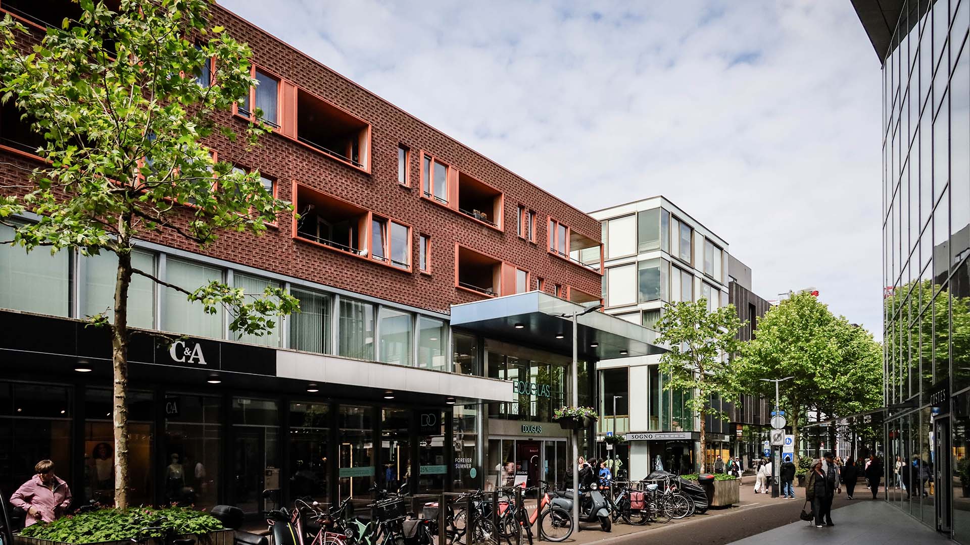 Winkelstraat met woningen boven de winkels, groene bomen, voetgangers en fietsen.