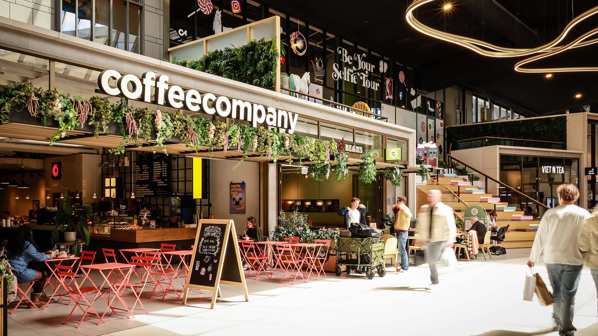 Binnenfoto winkelcentrum met winkel coffee company op voorgrond en winkelende mensen.
