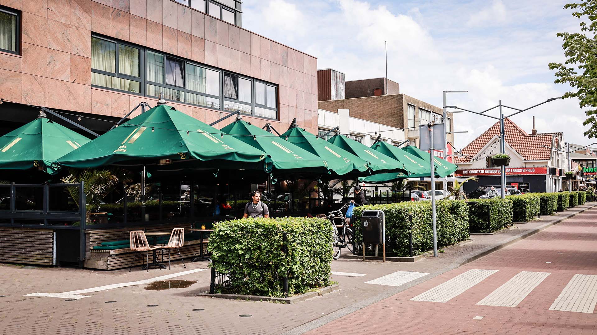 Horecaterras met groene parasols met kantoor erboven langs weg met zebrapad.