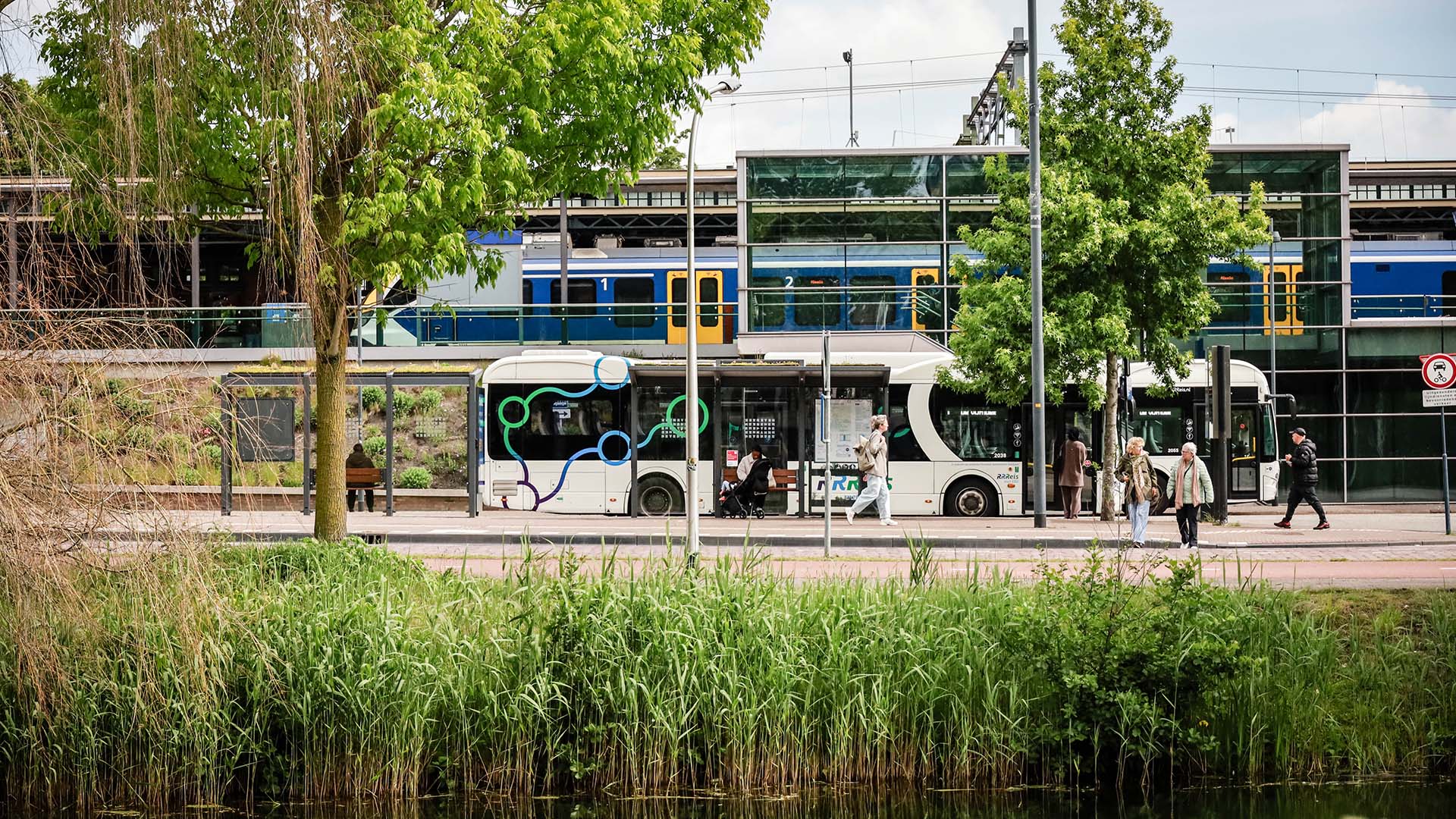 Groene bomen en riet op voorgrond, bus en trein met passagiers op station op achtergrond